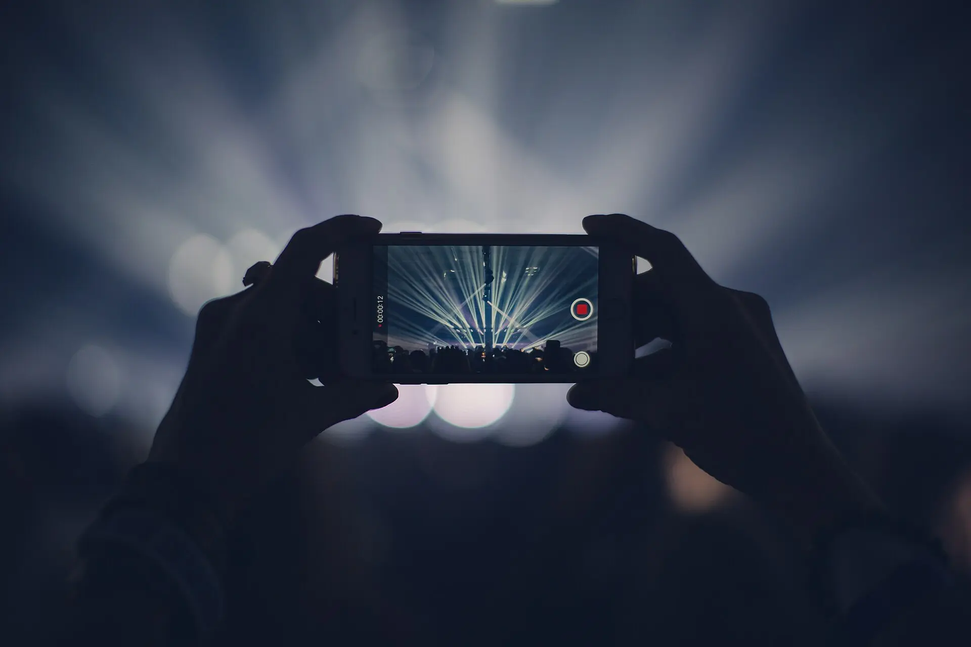 Silhouetted hands holding up a smartphone to film a concert stage