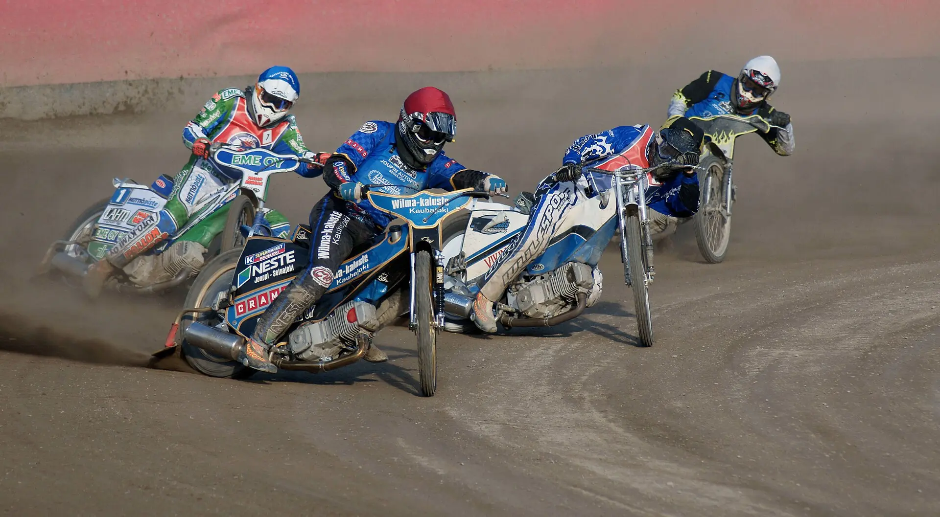 Four helmeted motorcycle riders leaning into a turn on a dirt race track